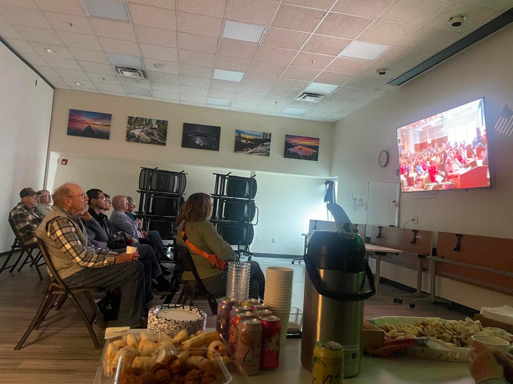A picture from the Free for All screening. While the documentary plays, a few rows of people watch the screen. In the foreground are snacks and drinks.