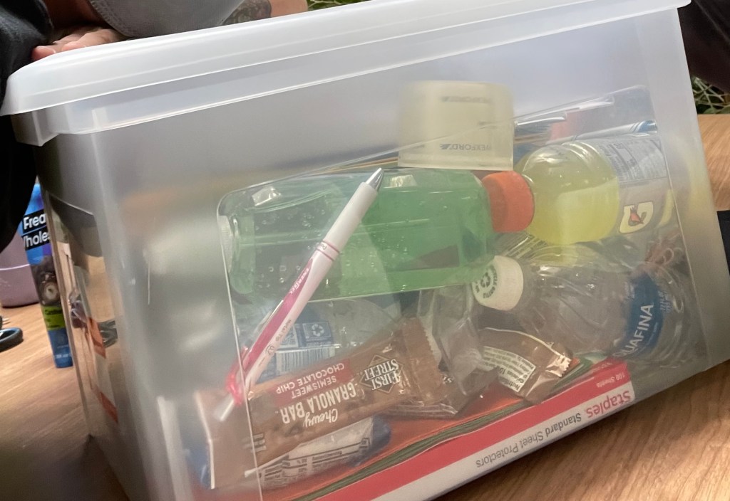 A clear plastic tub is on a wooden table. Inside is hand sanitizer, gatorade, bottled water, granola, snacks, and sheet protectors. There is clearly more in the tub as well.